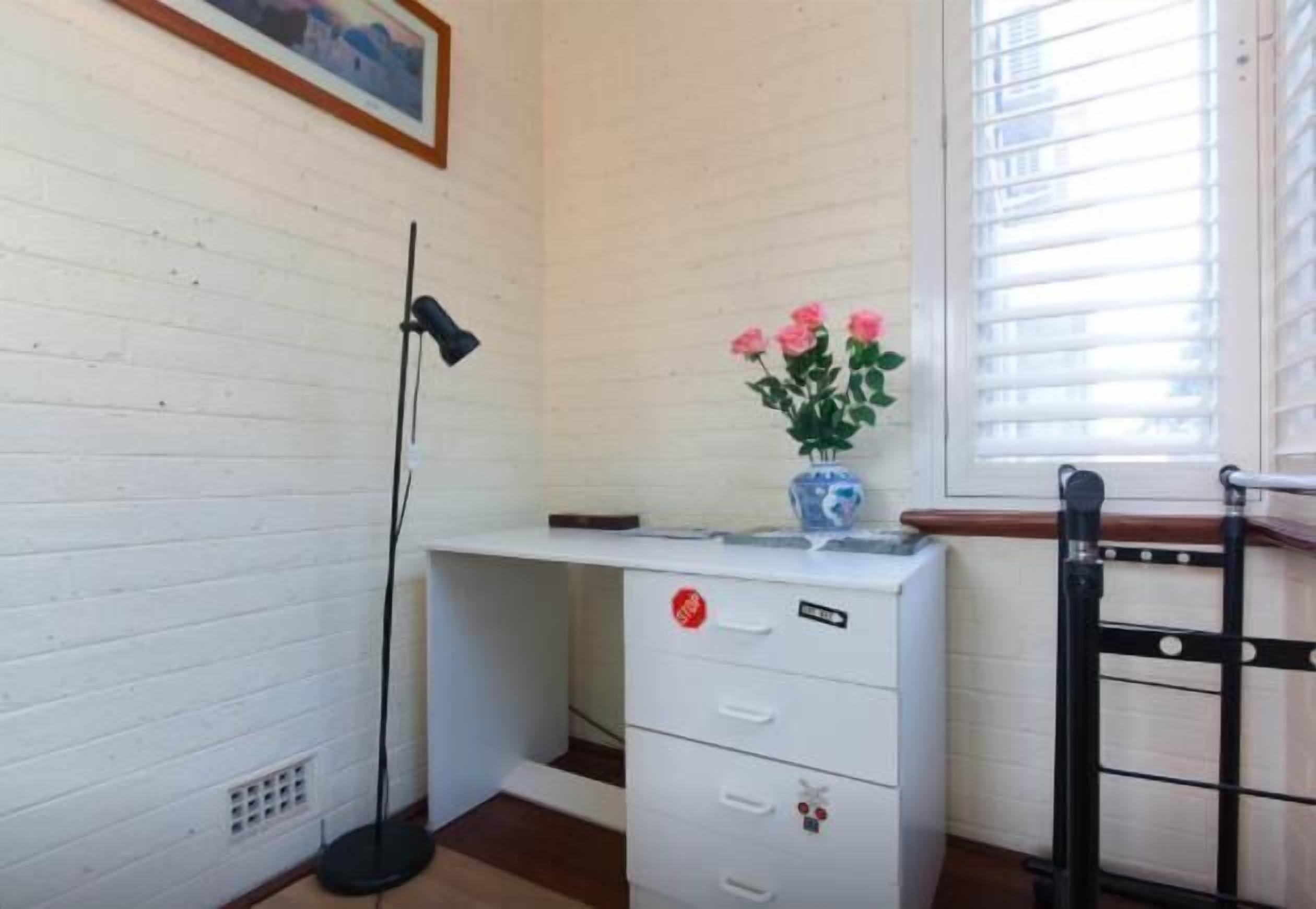 A white desk with drawers, a vase with pink flowers, a black floor lamp, and a window with blinds.