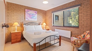 Un dormitorio con pared de ladrillo, cama con ropa de cama blanca, dos lámparas de noche, ventana con vista a árboles y una silla colorida.