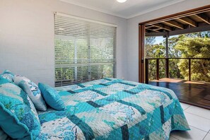 Une chambre avec un grand lit, une fenêtre avec des stores et un balcon donnant sur la verdure.