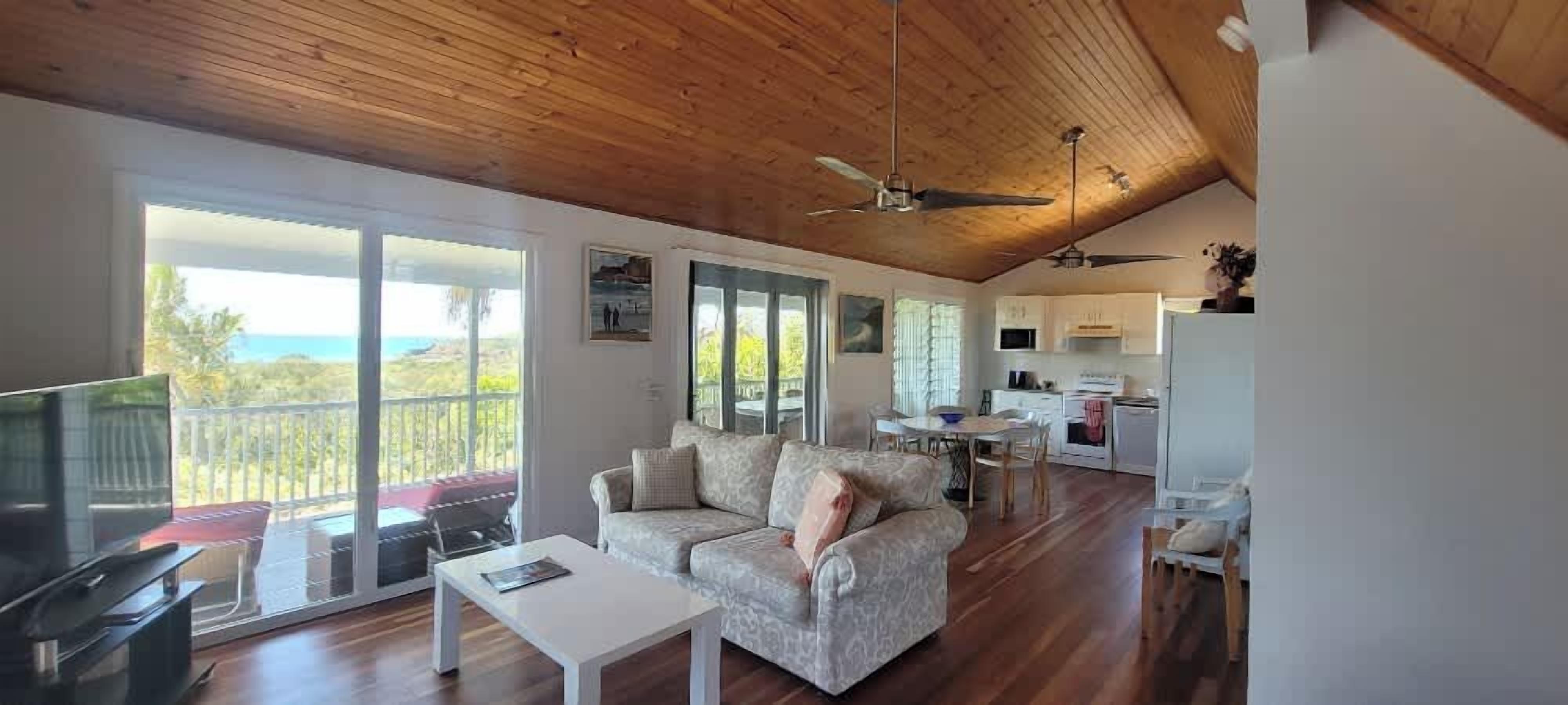 A spacious living area with a large sofa, a coffee table, and a TV. There is a balcony with a view of the ocean. The kitchen is visible in the background with white cabinets and a dining area.