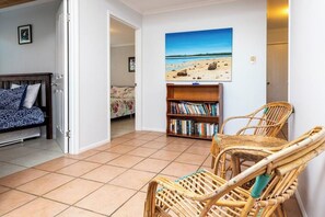 A living room with a bookshelf, two wicker chairs, a painting of a beach, and a bed with patterned bedding. - Arbour Rest Expansive Ocean Views (Agnes Water)