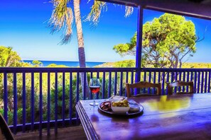 A wooden table with a glass of red wine and a plate of food, overlooking a beach with palm trees and a clear blue sky. - Arbour Rest Expansive Ocean Views (Agnes Water)
