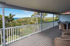 A covered outdoor area with a wooden deck, white railing, wicker furniture, and a view of the ocean and greenery. - Arbour Rest Expansive Ocean Views (Agnes Water)