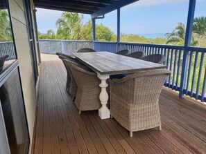 Une terrasse extérieure couverte, dotée d’une table en bois et de chaises en osier, qui offre une vue sur une plage bordée de palmiers.