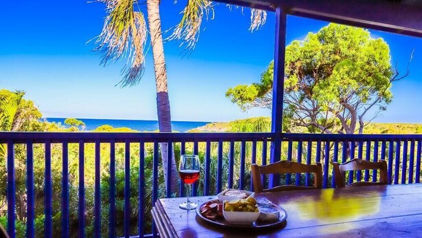Een houten tafel met een glas rode wijn en een bord eten, met uitzicht op een strand met palmbomen en een helderblauwe lucht.