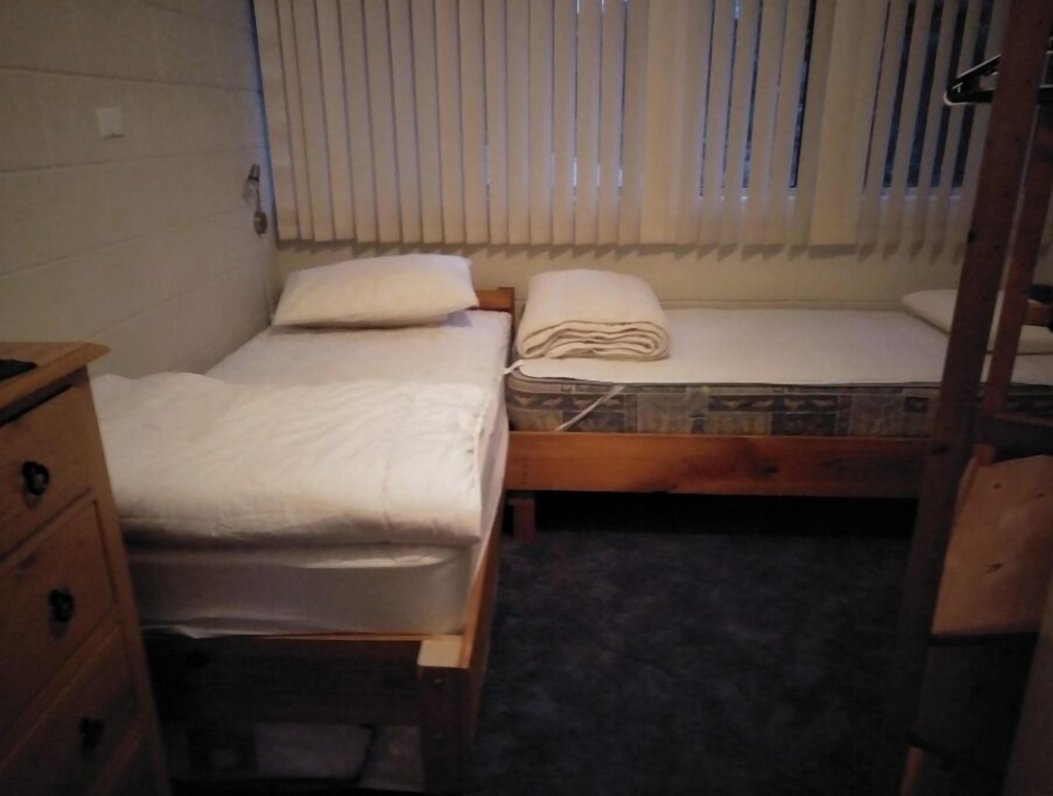 A small, dimly lit room with two beds, one with a wooden frame and the other with a metal frame. There is a wooden dresser with drawers and a small rug on the floor.