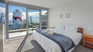 A modern hotel room with a large bed, a wooden nightstand, and a view of the sea through a sliding glass door.