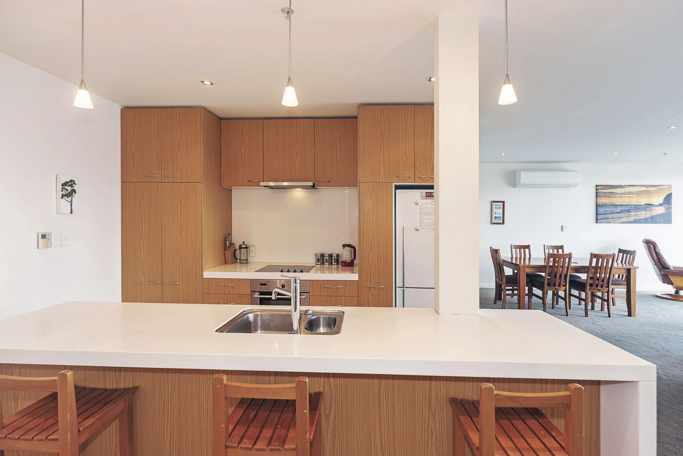 Une cuisine moderne avec des meubles en bois, un plan de travail blanc et un îlot central doté d’un évier. Trois chaises en bois sont placées devant l’îlot.