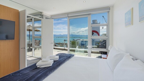 A modern hotel room with a large bed, a balcony with a view of the sea, and a glass door leading to an outdoor seating area.