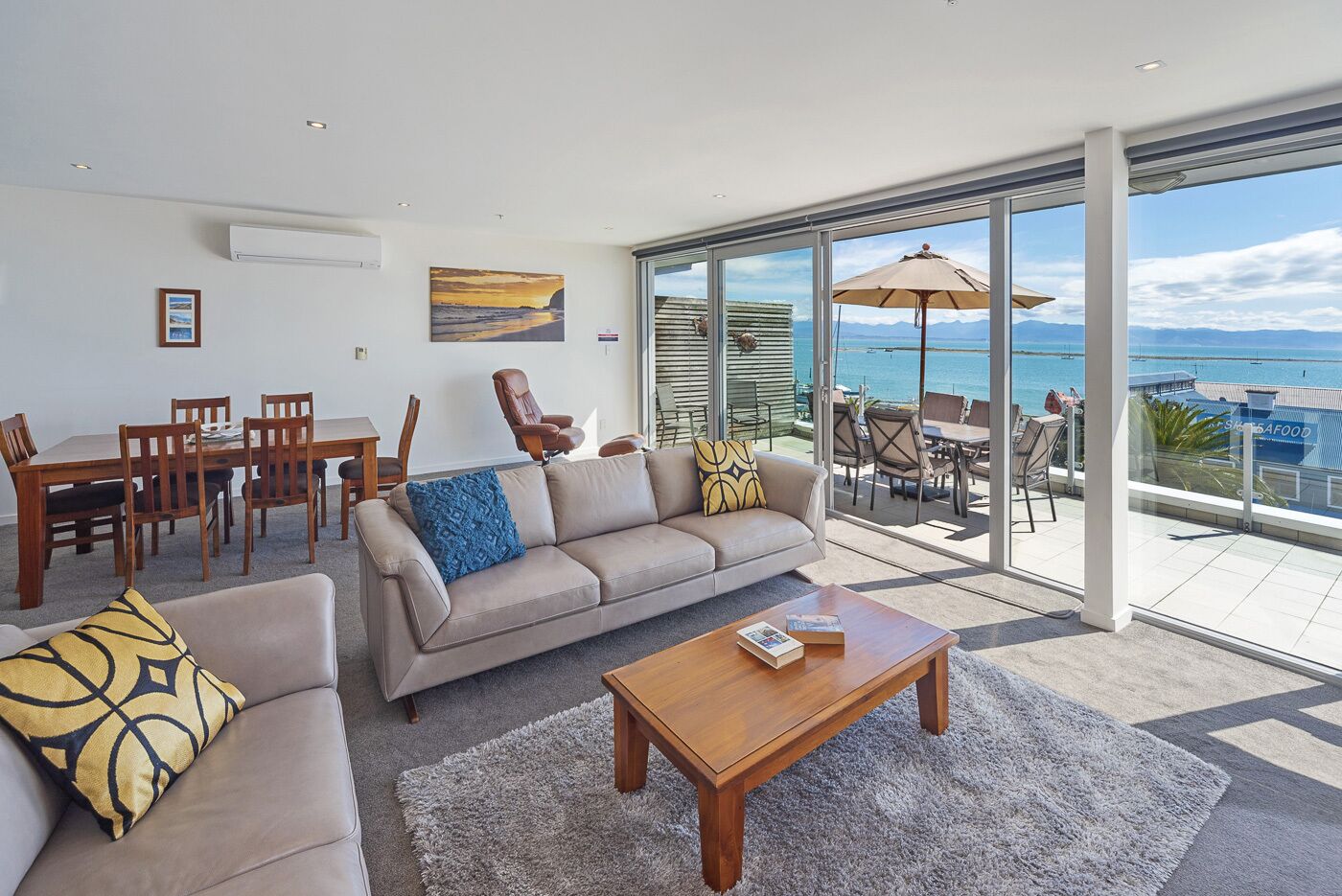 Een ruime woonkamer met een grote bank, een salontafel en een eethoek. Een groot raam biedt uitzicht op de zee en er is een balkon met tuinmeubilair.