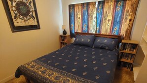 Ein Schlafzimmer mit einem großen Bett, einem Kopfteil mit bunter Vorhang, einem kleinen Nachttisch mit Lampe und einem Bücherregal.