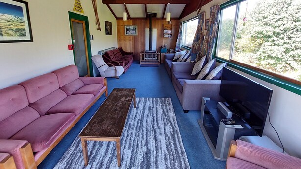 Ein gemütliches Zimmer mit einer Couch, einem kleinen Tisch und einem Fernseher. Holzbalken sind an der Decke und es gibt ein Fenster mit Blick auf Bäume.