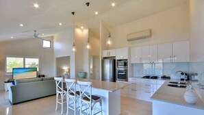 Una cocina moderna con armarios blancos, una isla central, taburetes de bar y una zona de comedor con sofá y televisor.