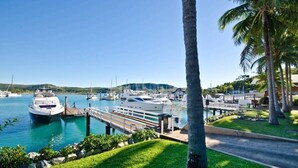 Un puerto deportivo con barcos amarrados, una zona verde con palmeras y un cielo azul despejado.