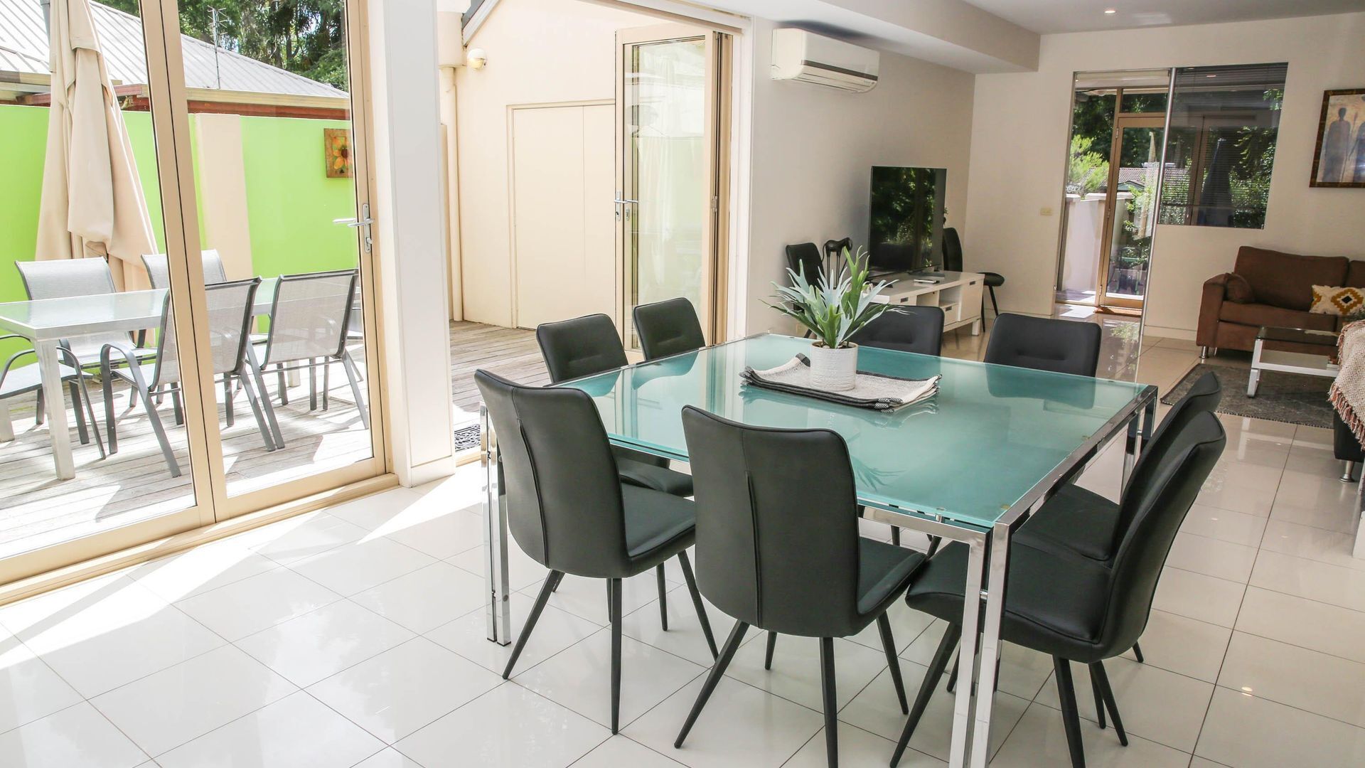 A modern dining area with a glass table and black chairs, a green wall, and a sliding glass door leading to an outdoor patio.