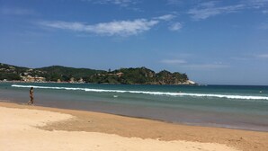 A beach with a person walking along the shore, waves gently crashing onto the sand, and a distant view of a hilly landscape with buildings. - Wonderful house in Geribá, 4 suites, balcony and gourmet area. (BÚZIOS)