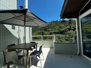 Eine Terrasse im Freien mit Tisch und Stühlen, einem großen Sonnenschirm und Blick auf einen bewaldeten Hügel.