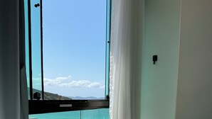 A hotel room with a large window overlooking a beach with umbrellas and people. - Loft with sea view. (apartamento 501 prainha Arraial do CABO)