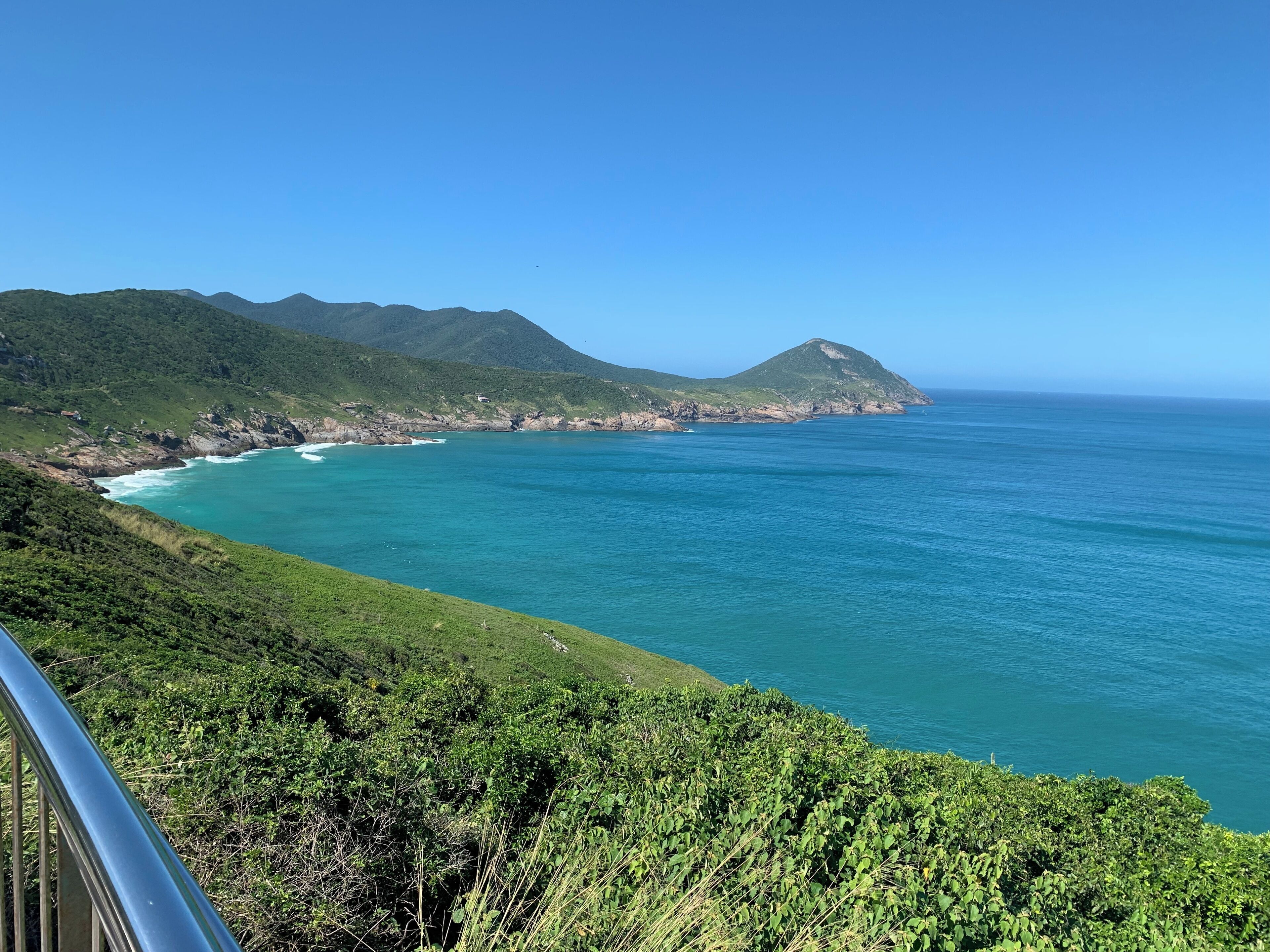 A coastal landscape with a clear view of the sea, green hills, and a distant mountain range.