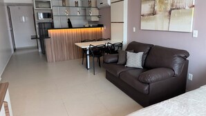 A modern living room with a brown leather sofa, a dining table with chairs, and a kitchen area with a microwave and a coffee machine. - Loft with sea view. (apartamento 501 prainha Arraial do CABO)