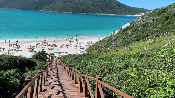 A wooden staircase leading to a beach with turquoise water and green hills in the background. - Loft with sea view. (apartamento 501 prainha Arraial do CABO)