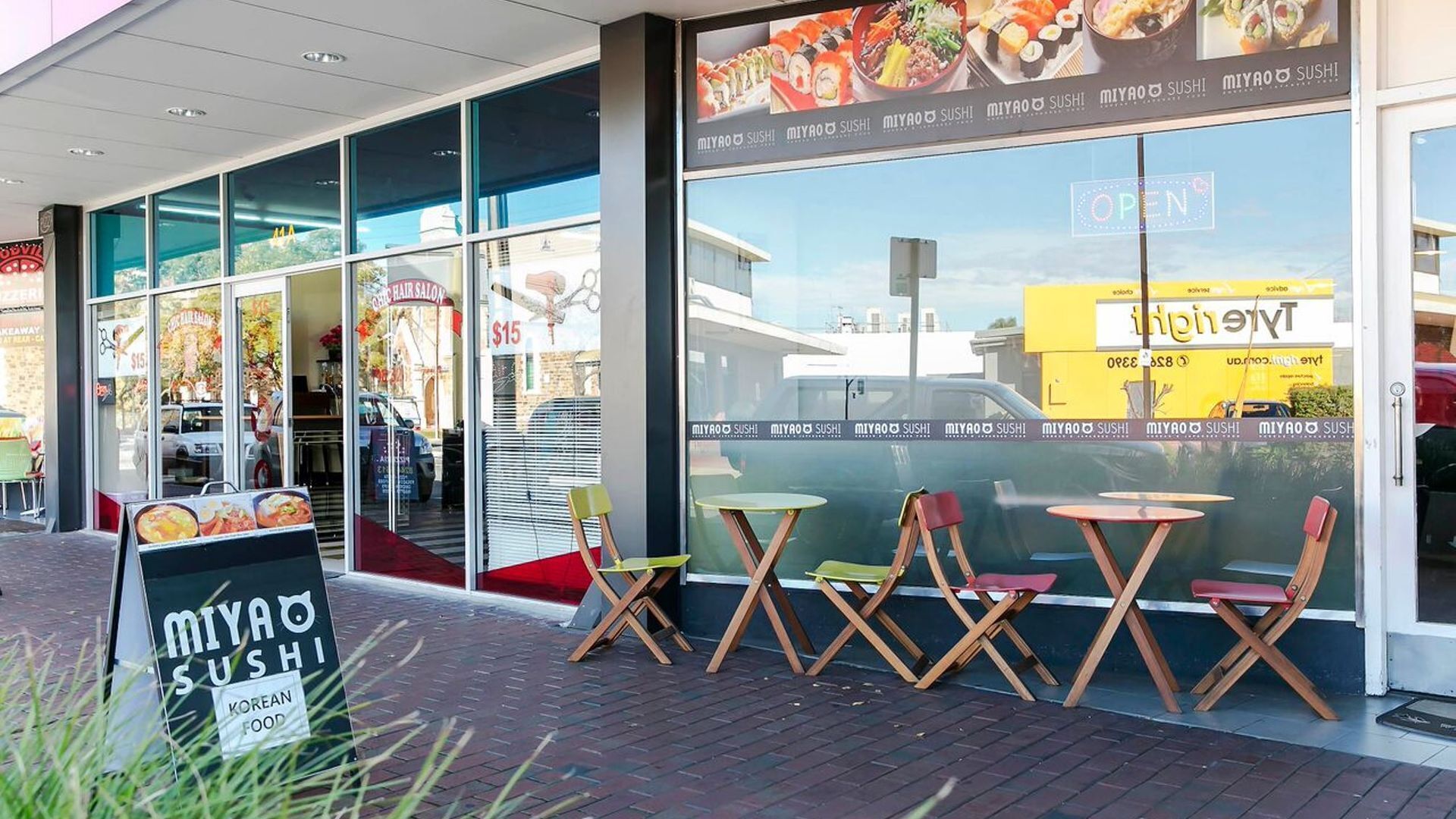 A storefront with a sign for 'MIYAO SUSHI' and a 'OPEN' sign. There are outdoor seating areas with tables and chairs.