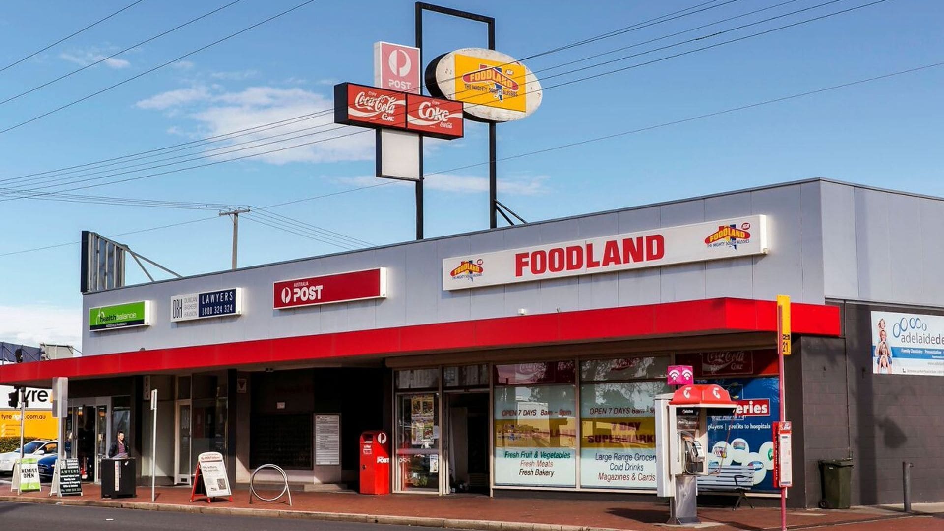 A FOODLAND supermarket with a red and white facade, a Coca-Cola sign, and a sign for Australia Post.