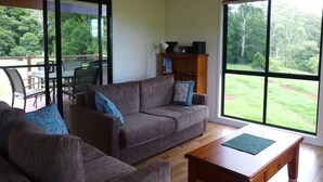 A living room with a grey sofa, a wooden coffee table, and a bookshelf. There is a glass door leading to an outdoor area with a table and chairs.