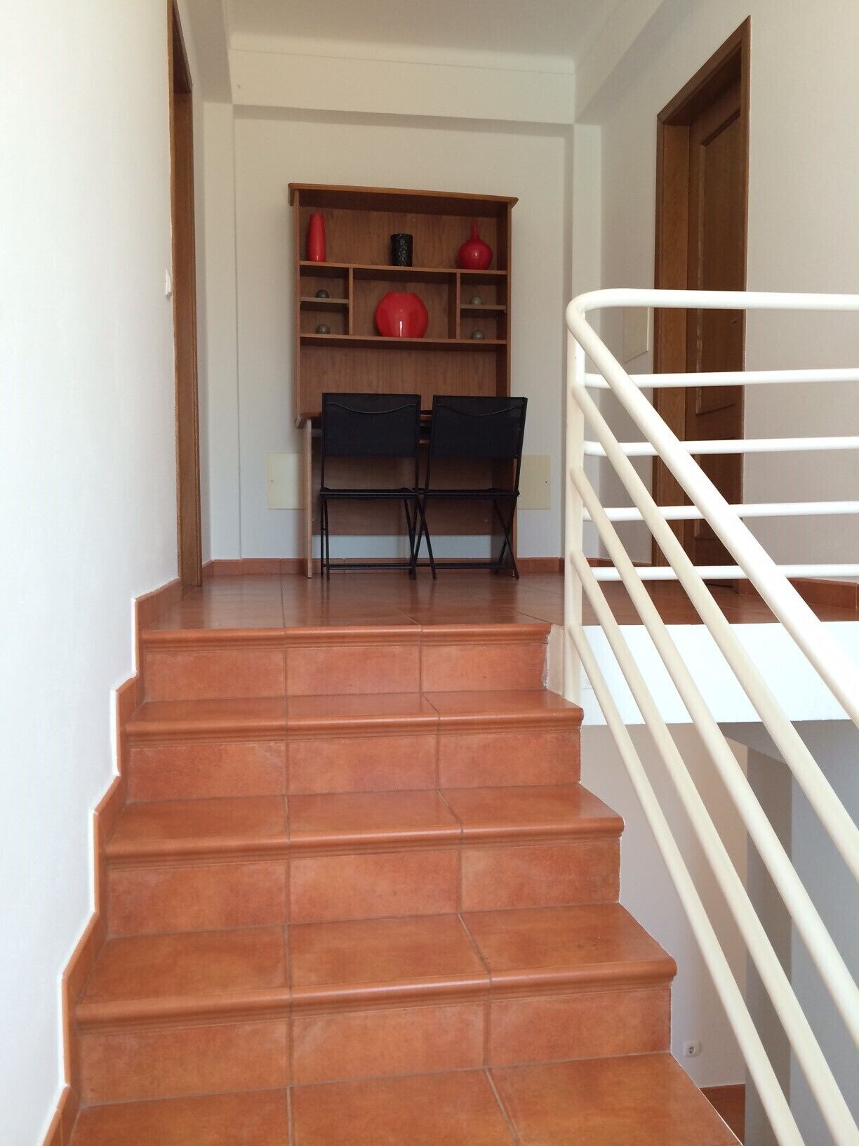 Una escalera con barandilla blanca que conduce a una plataforma con un estante de madera y dos sillas negras.