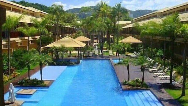Zwembadgedeelte van een resort met een lang zwembad, palmbomen en ligbedden.