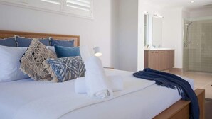 A bedroom with a bed, pillows, a bedside table, a mirror, and a bathroom with a shower. - Green House Pet-Friendly Beach House (Mount Coolum)
