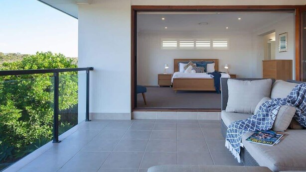 A modern bedroom with a large bed, bedside tables, and a balcony with a glass railing. - Green House Pet-Friendly Beach House (Mount Coolum)