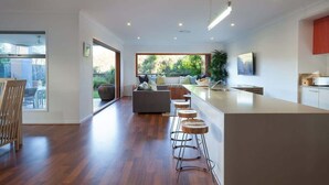 A modern kitchen with a central island, bar stools, and a dining area with a wooden table and chairs. - Green House Pet-Friendly Beach House (Mount Coolum)