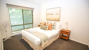 Une chambre avec un grand lit, une fenêtre avec des stores, une table de chevet avec une lampe et une œuvre d’art accrochée au mur.