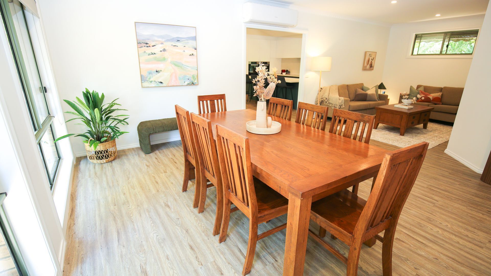 A dining area with a wooden table and chairs, a potted plant, and a painting on the wall.