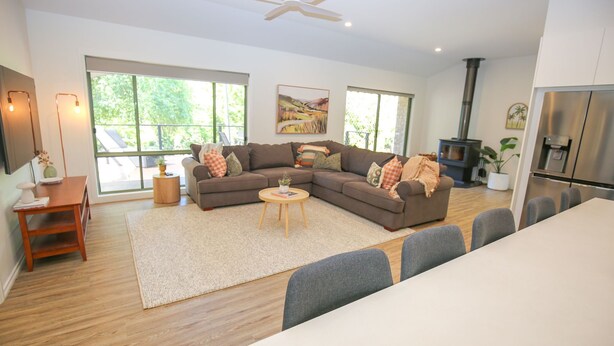 Un salon moderne avec un canapé d’angle, une table basse ronde et un coin repas avec des chaises grises. Une grande fenêtre laisse entrer la lumière naturelle et offre une vue sur la verdure à l’extérieur.
