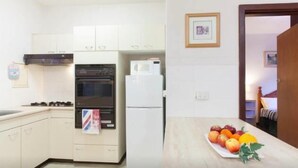 A kitchen with white cabinets, a black oven, and a white refrigerator. There is a framed picture on the wall and a fruit plate on the counter.