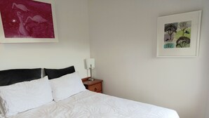 En snyggt bäddad säng med vita sängkläder och ett blommönster, ett träbord vid sängen med en lampa och två inramade konstverk på väggen.