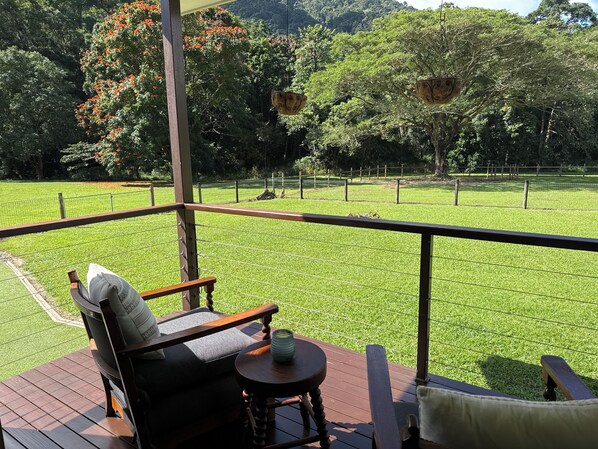 A wooden deck with a chair and a small table, overlooking a fenced grassy area with trees and hanging planters.