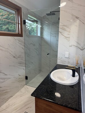 Un baño moderno con cabina de ducha de vidrio, pared revestida de mármol, encimera de granito negro y lavabo blanco.