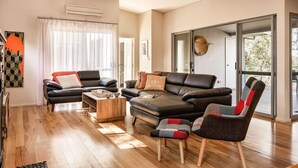 Ein modernes Wohnzimmer mit einer schwarzen Ledersofa, einem Holztisch und einem farbenfrohen Sessel. Ein großes Fenster mit durchsichtigen Vorhängen, ein Deckenventilator und eine Wandklimaanlage sind vorhanden.