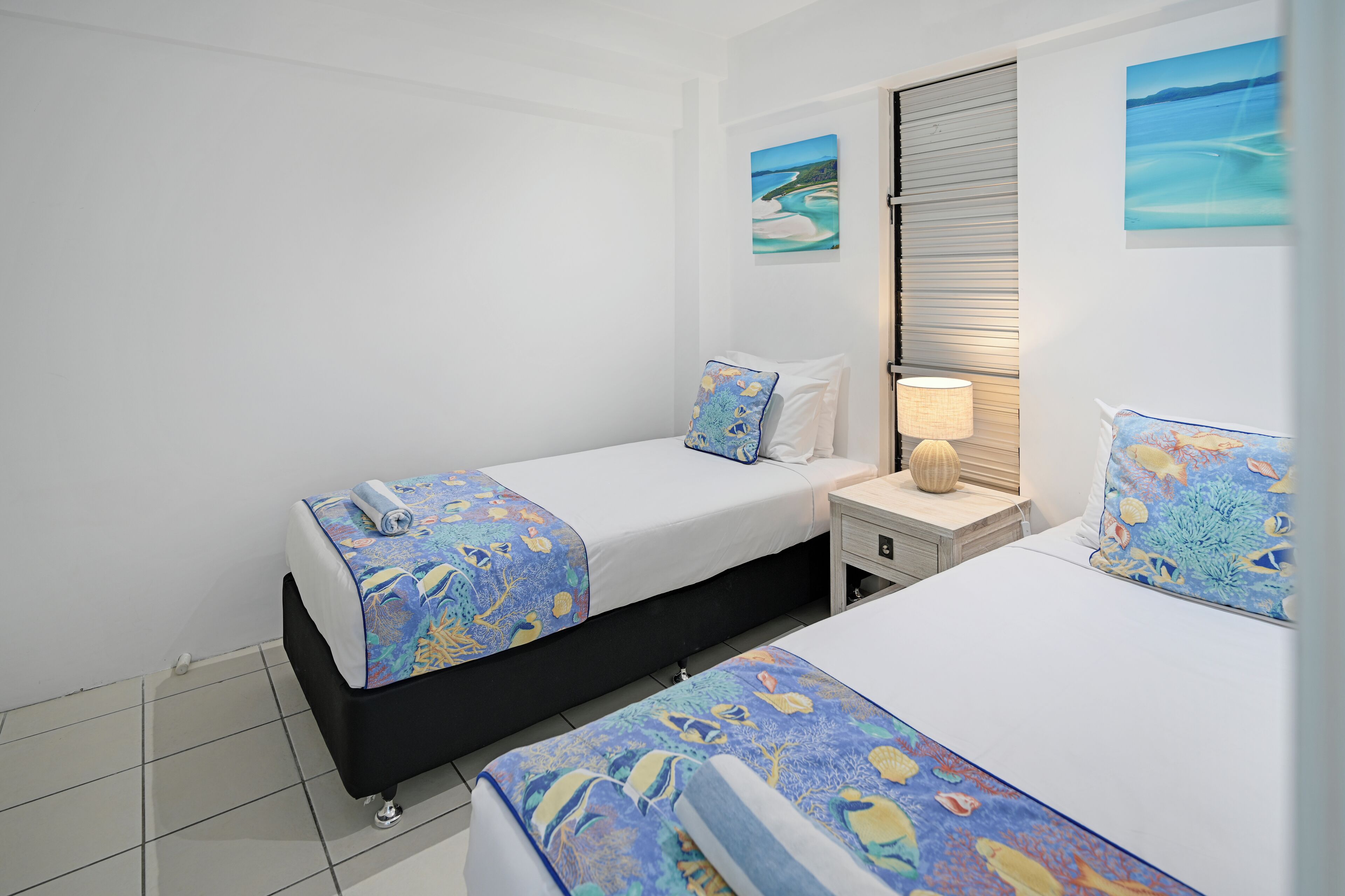 Une chambre d’hôtel avec deux lits, chacun recouvert d’une couvre-lit à motifs bleus et jaunes, agrémentée de coussins blancs et d’une petite table de chevet avec une lampe.