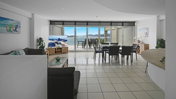 A modern living room with a large TV, a dining area with chairs, and a view of the sea through large windows. - Ocean View Villa - 02  (Hamilton Island)
