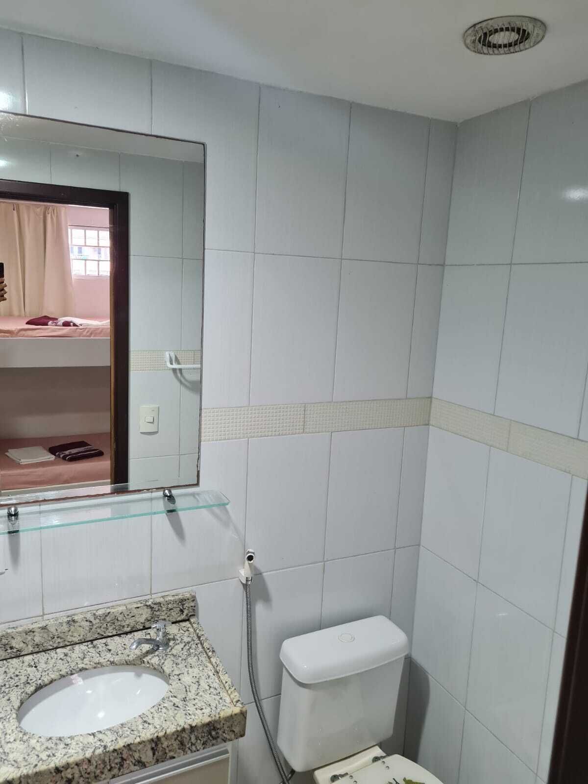 Une salle de bain avec un mur recouvert de carreaux blancs, une étagère en verre au-dessus du lavabo, des toilettes blanches et un miroir qui reflète un lit près d’une fenêtre.