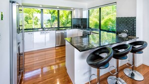 Une cuisine moderne dotée d’un grand îlot central, de tabourets de bar et offrant une vue sur la verdure grâce à de grandes fenêtres.