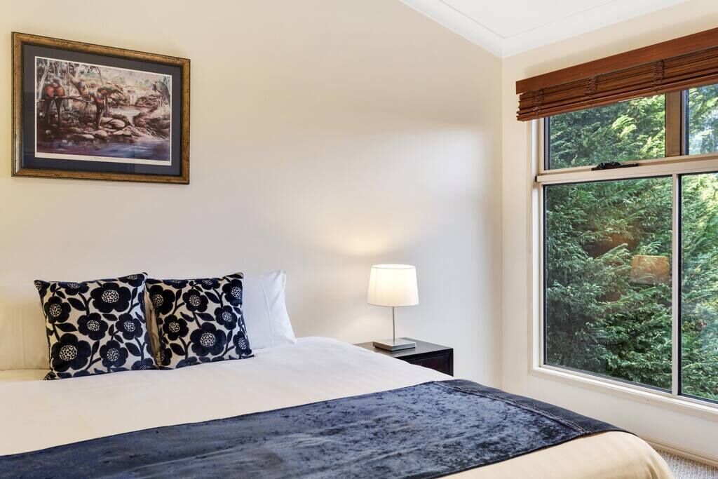 A hotel room with a bed, a nightstand with a lamp, a framed picture on the wall, and a window with a view of trees.