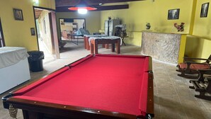 Una mesa de billar en una habitación con paredes amarillas, techo de madera y una pared decorativa de piedra. Se ven sillas y una pequeña mesa al fondo.