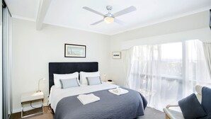Una habitación de hotel con cama, escritorio, silla, ventilador de techo y ventana con cortinas translúcidas.