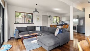 A modern living room with a grey sectional sofa, a wooden coffee table, and a blue rug. A kitchen with white cabinets and stainless steel appliances is visible in the background. - Cosy Corner | Margaret River (Margaret River)