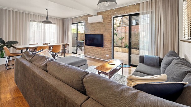 A modern living room with a large sofa, a coffee table, and a TV mounted on a brick wall. There is a dining area with a table and chairs, and a sliding glass door leading to an outdoor space. - Cosy Corner | Margaret River (Margaret River)